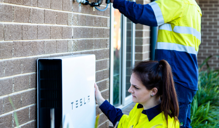 two battery installers are wearing yellow blue tops and installing battery to a house